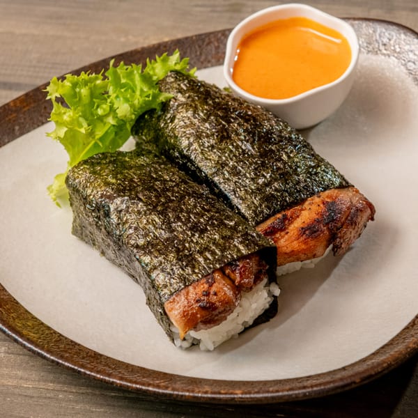 Two Spam Musubi with rice and seaweed, garnished with lettuce and a side of orange sauce.